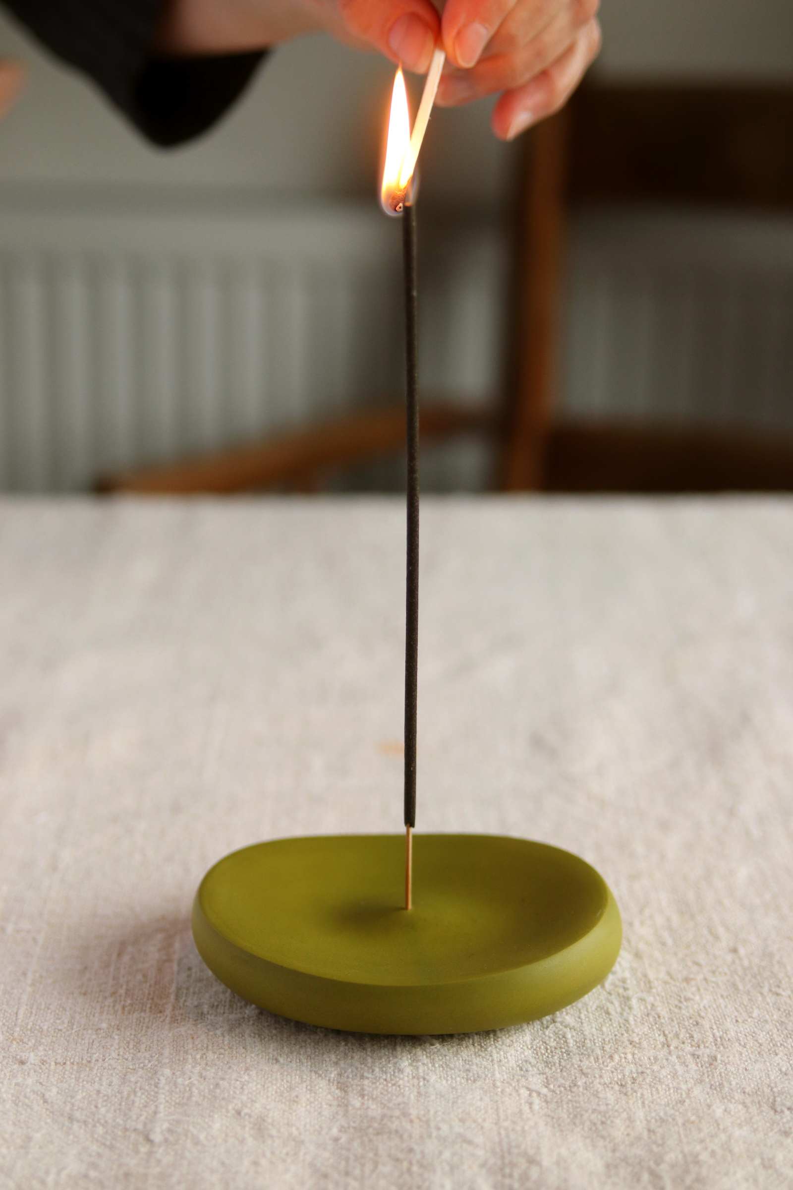 A hand lighting an incense stick on a round olive green incense holder made of jesmonite, placed on a white surface.